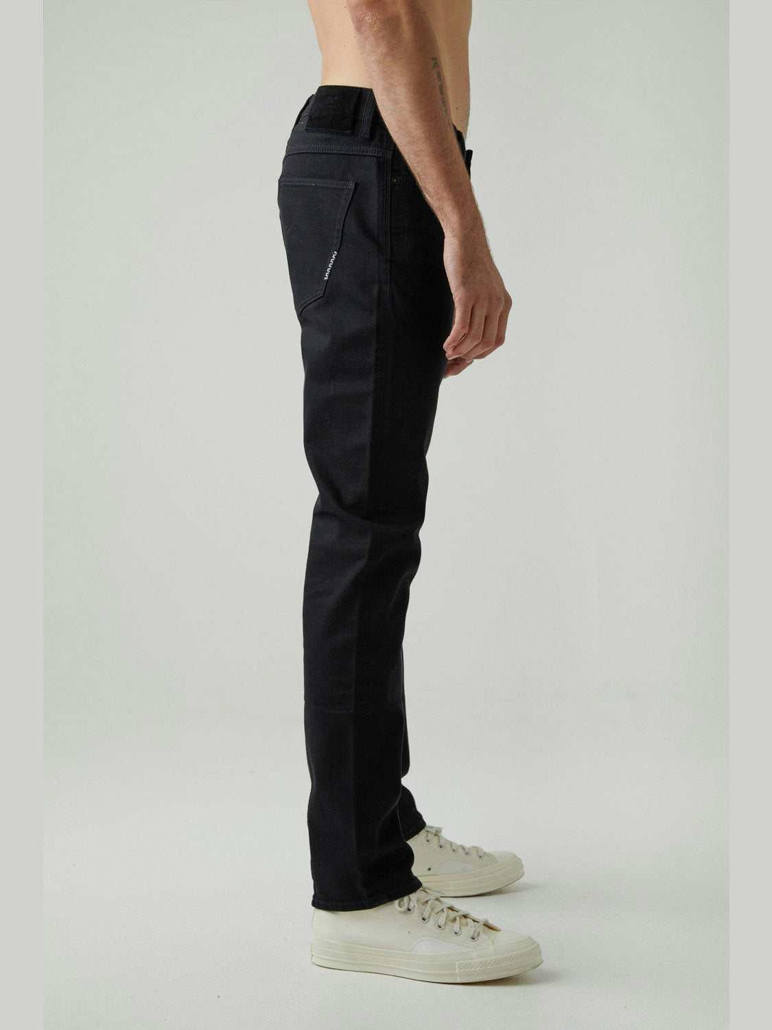 A person stands sideways against a plain light background, wearing Neuw Lou Slim Forever Black jeans with comfort stretch and white sneakers. Only their lower torso and legs are visible; they are shirtless.