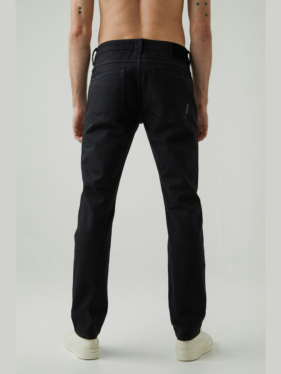 A shirtless person faces away from the camera, wearing Neuw's Lou Slim Forever Black jeans and white sneakers against a plain light background.