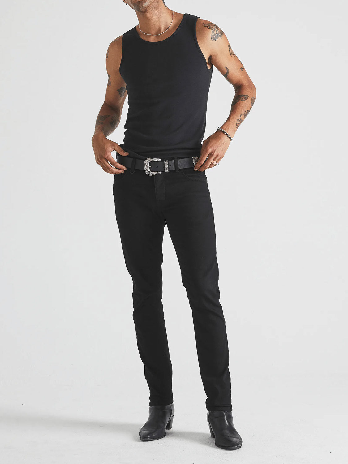 A person stands against a white background wearing Neuw's Lou Slim Forever Black jeans, a black tank top, a black belt with silver buckle, and black boots. Their arms are tattooed and they are adjusting the belt.