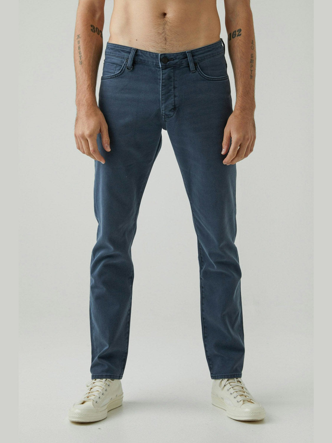 A man faces forward, wearing Neuw’s Lou Slim Liberte jeans in vintage grey stretch denim with white sneakers. The photo, cropped at the shoulders, showcases his bare torso and limbs against a plain background.