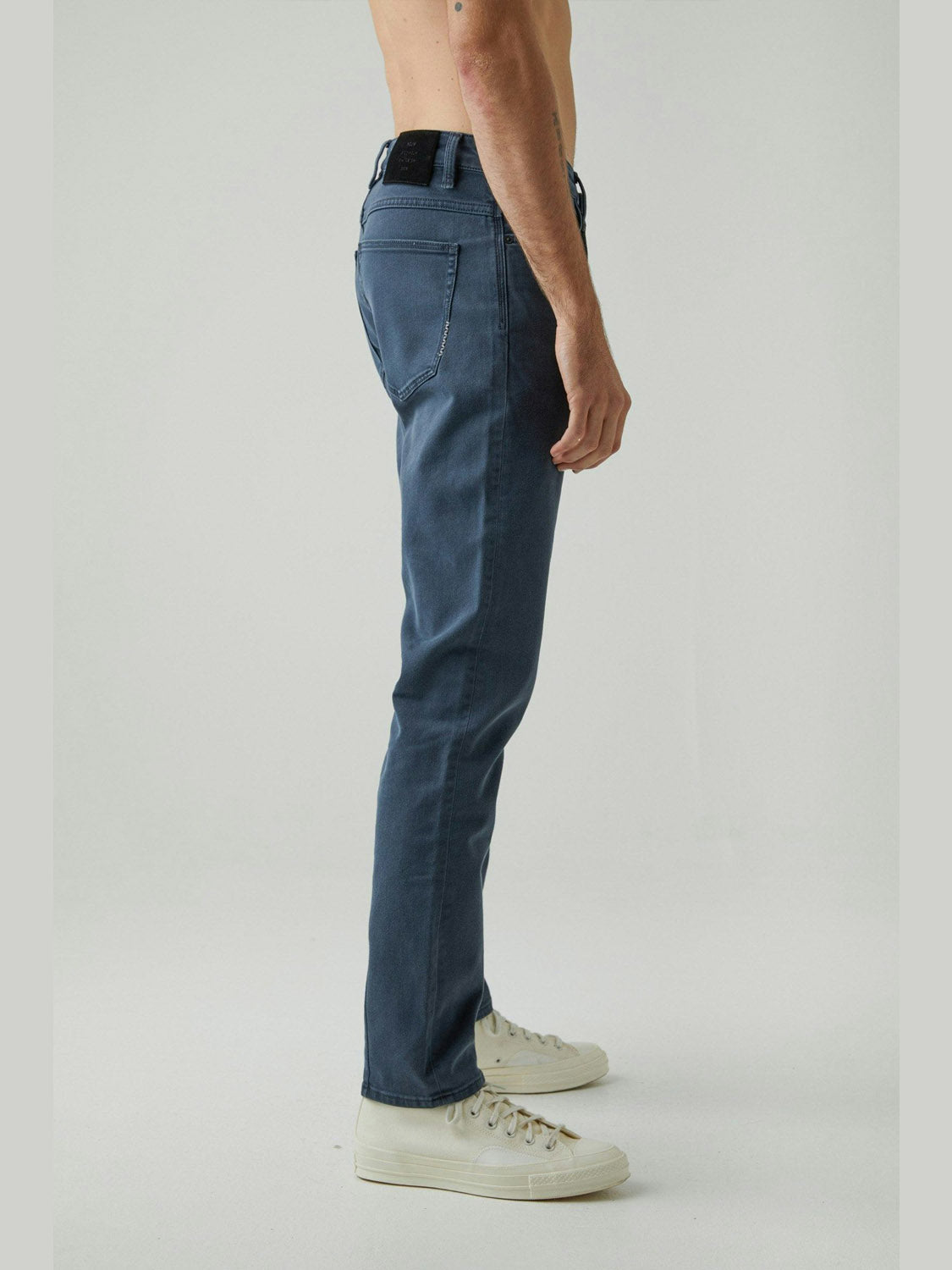A person stands in profile wearing Neuw Lou Slim Liberte jeans in a vintage grey wash and white sneakers, bare torso, against a plain light background. Made from premium stretch denim, these jeans deliver both style and comfort.
