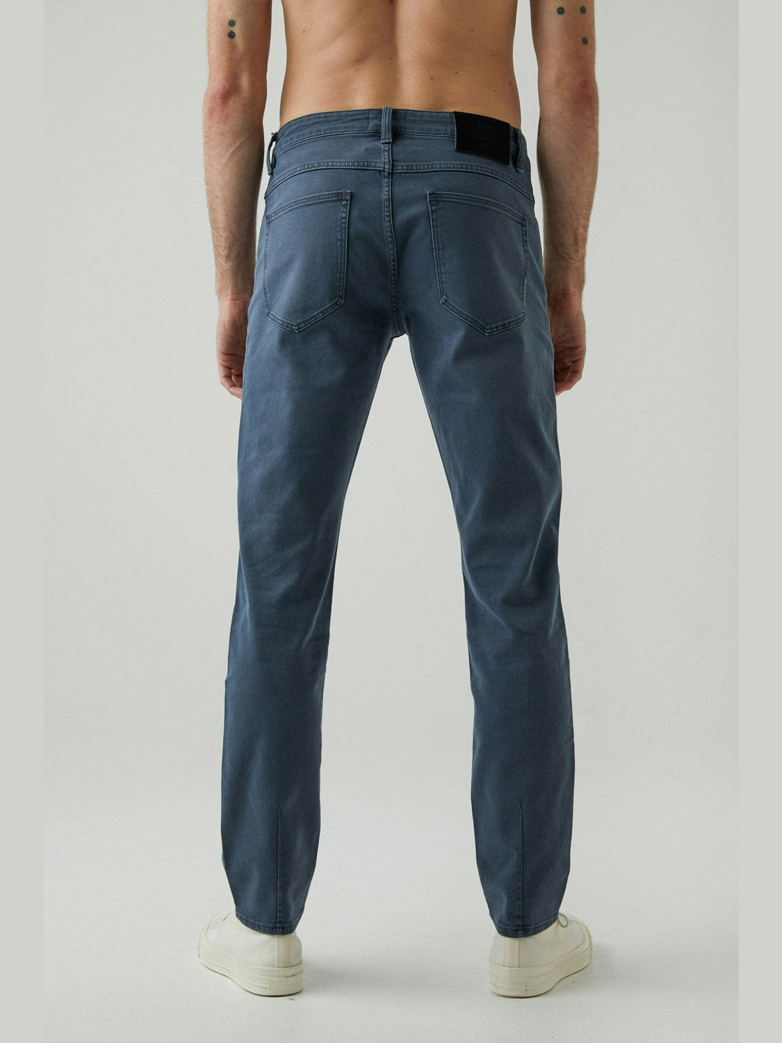 A shirtless person stands facing away, wearing Neuw Lou Slim Liberte jeans in vintage grey wash with white sneakers, shown from the waist down against a plain light background.