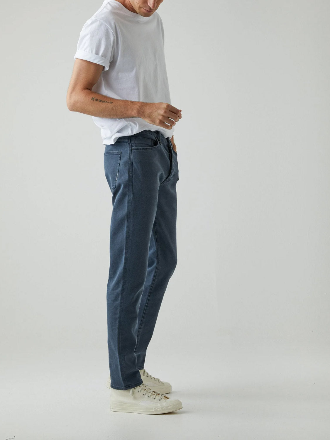 A person wearing a white t-shirt, Neuw Lou Slim Liberte jeans in vintage grey wash, and white sneakers stands against a plain, light background, with the upper part of their face out of frame.