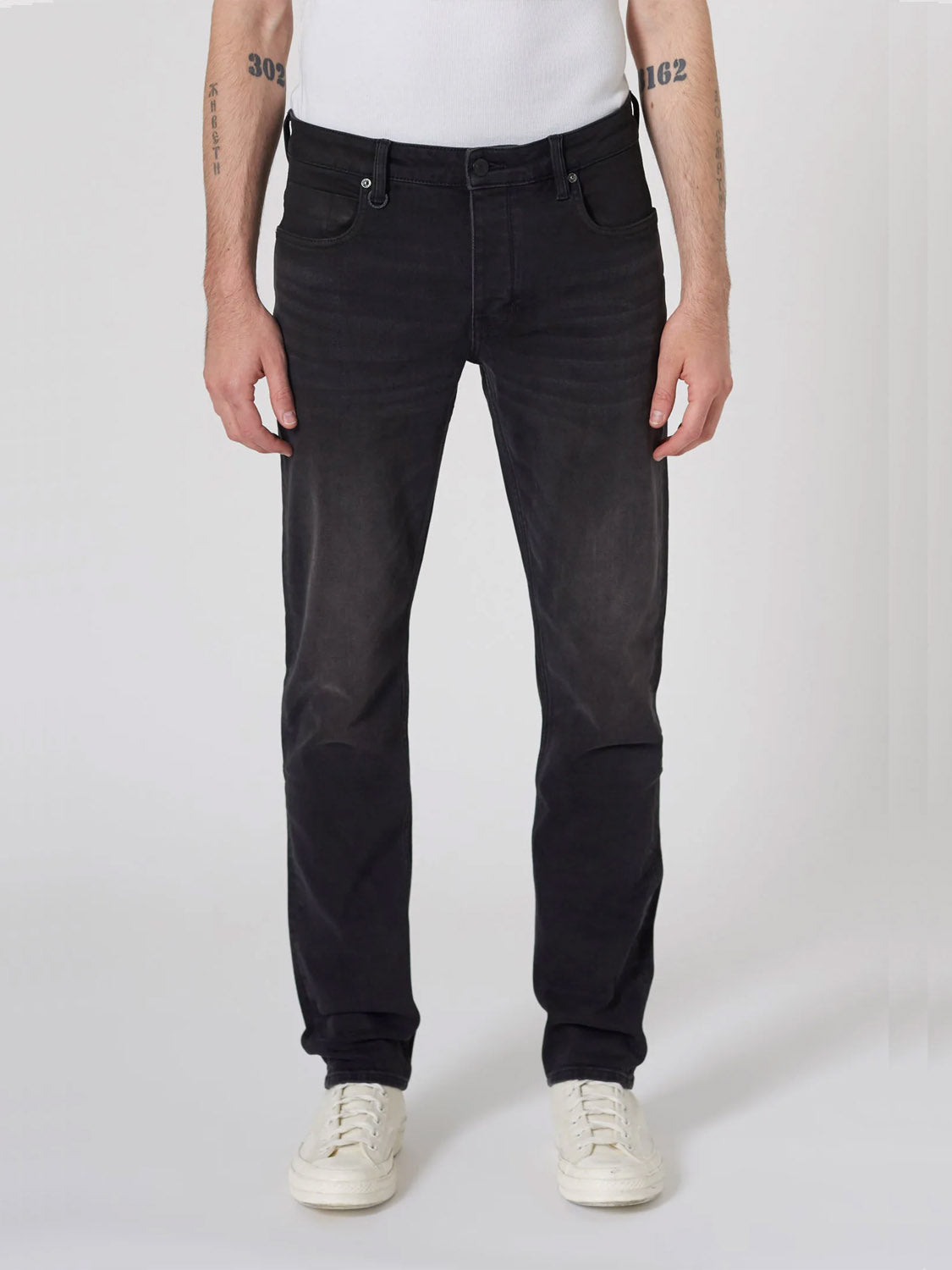 A person wears Neuw's Lou Slim Reel jeans made from premium stretch fabric, paired with a white sleeveless shirt and white sneakers, standing against a plain white background. Only the lower body and arms are visible.