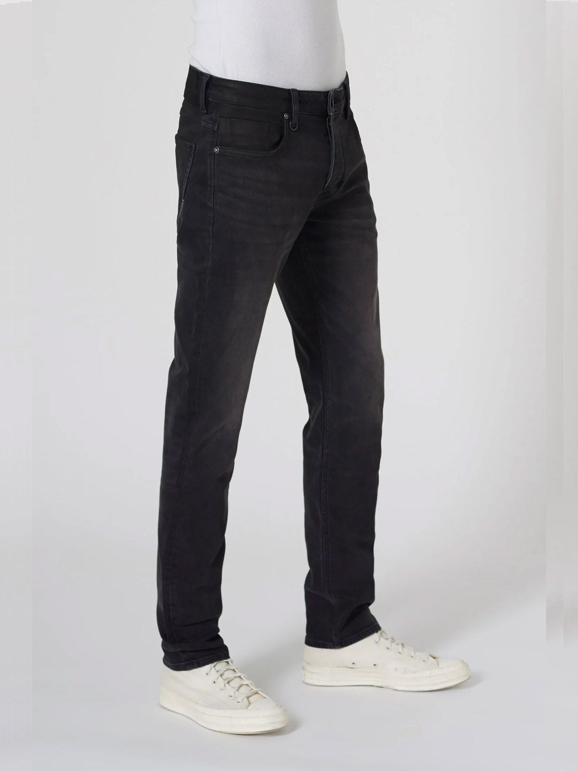A person wearing Neuw Lou Slim Reel jeans made from premium stretch fabric and white sneakers stands against a plain light gray background, with only the lower half of their body visible.