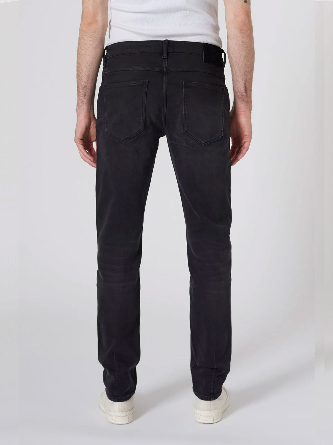 Someone models the Neuw Lou Slim Reel jeans in organic black denim, paired with a white tank top and white sneakers, standing with their back to the camera against a plain light background.
