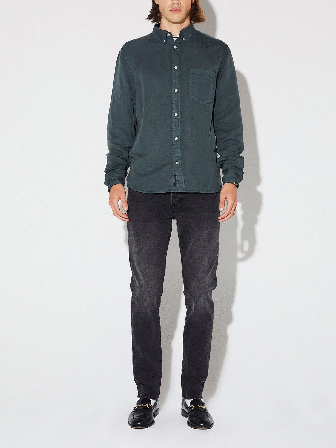 A person stands against a plain background wearing a dark green button-up shirt, Neuw's Lou Slim Reel slim fit jeans in organic black denim, and black loafers, facing forward with arms relaxed by their sides.
