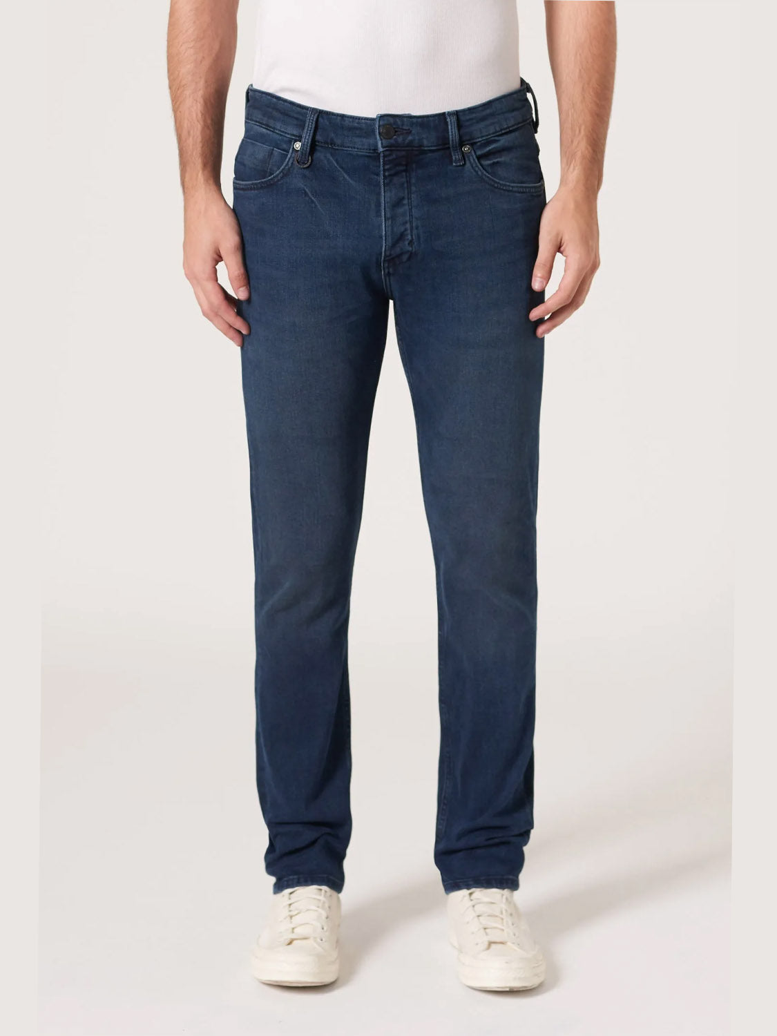 A person wearing Neuw Lou Slim Silent Water indigo denim jeans, a white shirt, and white sneakers stands against a plain light background. The image is cropped at the shoulders.