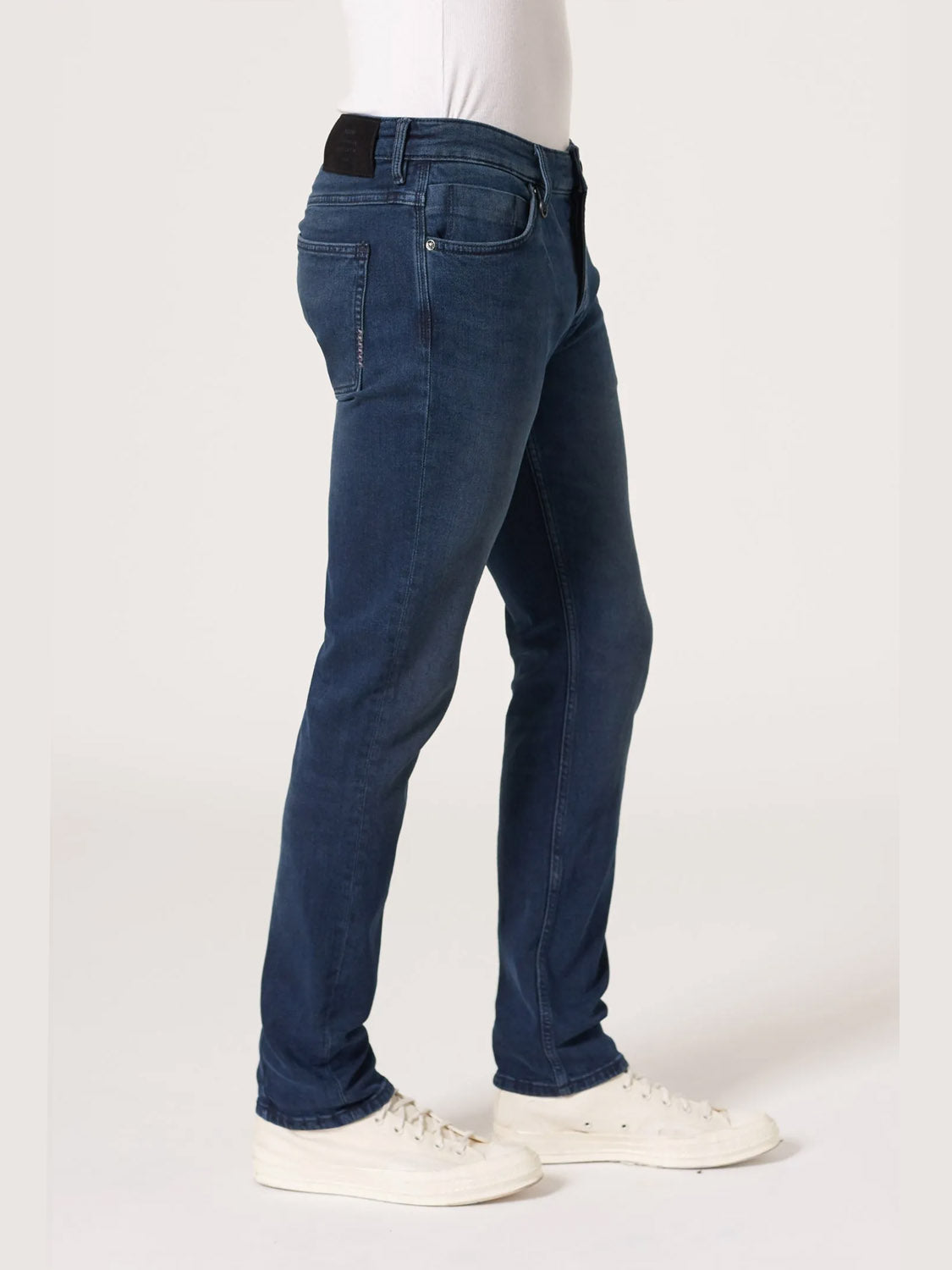 A person wears Neuw’s Lou Slim Silent Water indigo jeans, crafted from organic cotton, with white sneakers. Shown from the waist down, they stand sideways against a plain light background.
