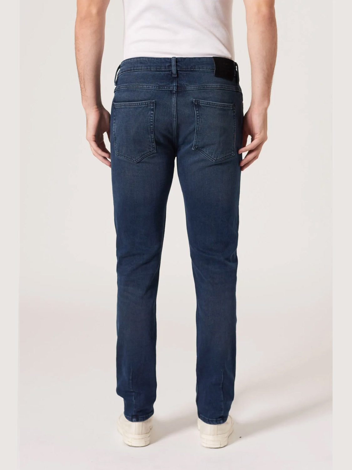 A person is shown from the back against a plain light background, wearing Neuw's Lou Slim Silent Water indigo denim jeans, a white shirt, and white sneakers.