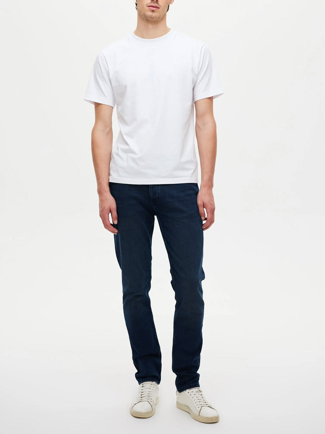 A person stands against a plain white background, wearing Neuw's Lou Slim Silent Water indigo denim jeans with a white short-sleeve t-shirt and white sneakers. Their arms are relaxed at their sides, and their face is partially visible.