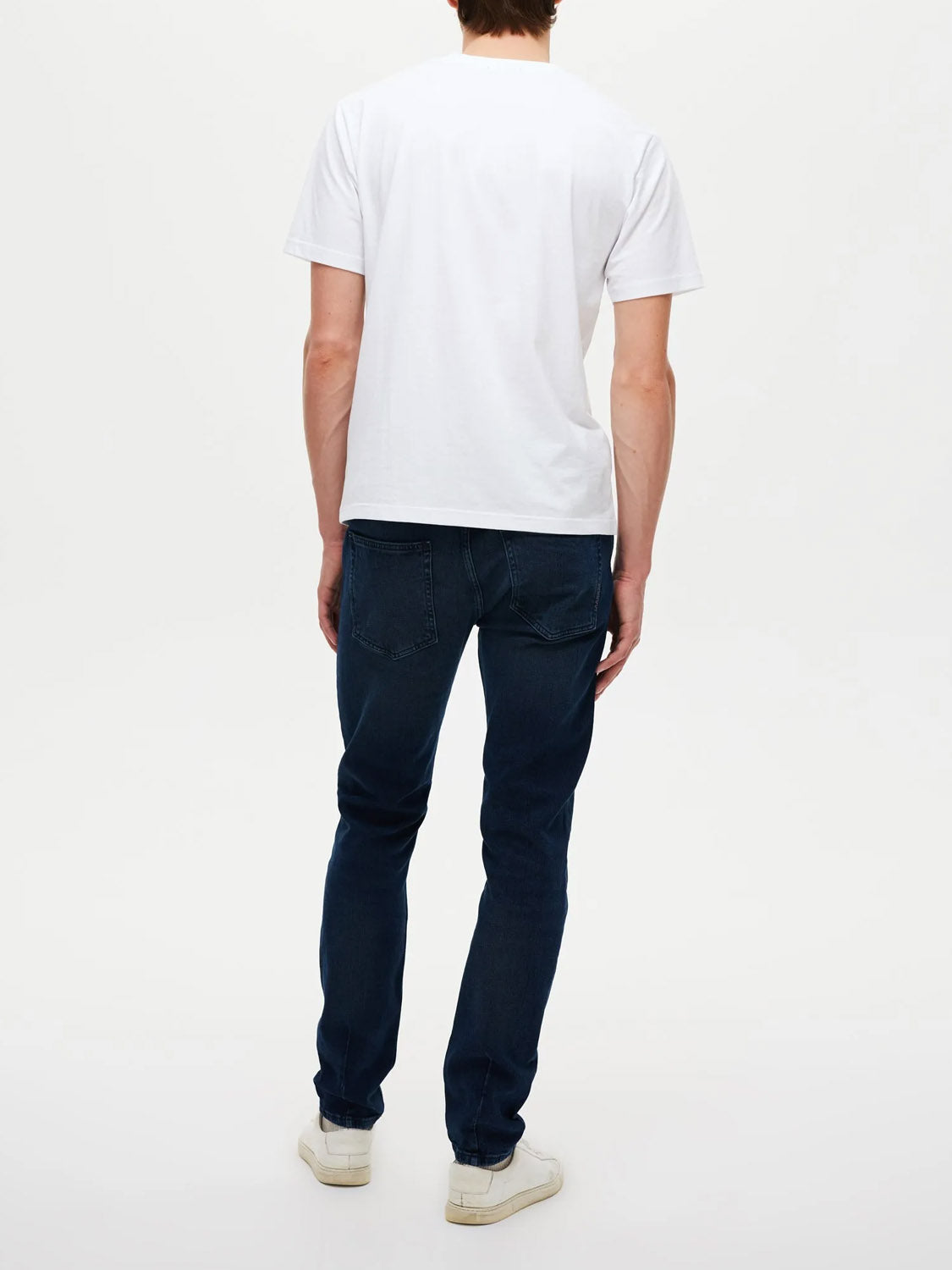 A person stands with their back to the camera, wearing Neuw's Lou Slim Silent Water indigo denim jeans, a plain white T-shirt, and white sneakers against a plain white background.