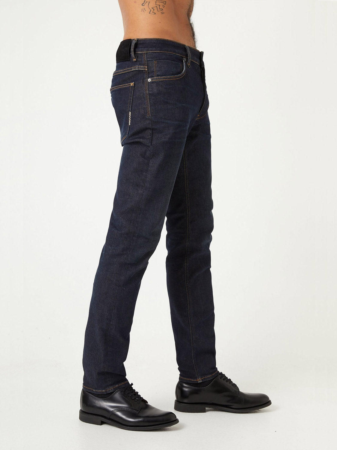 A person poses sideways in ID Menswear Lou Slim - Typecast jeans and black dress shoes, with a visible left-side tattoo and no shirt, against a plain white background.