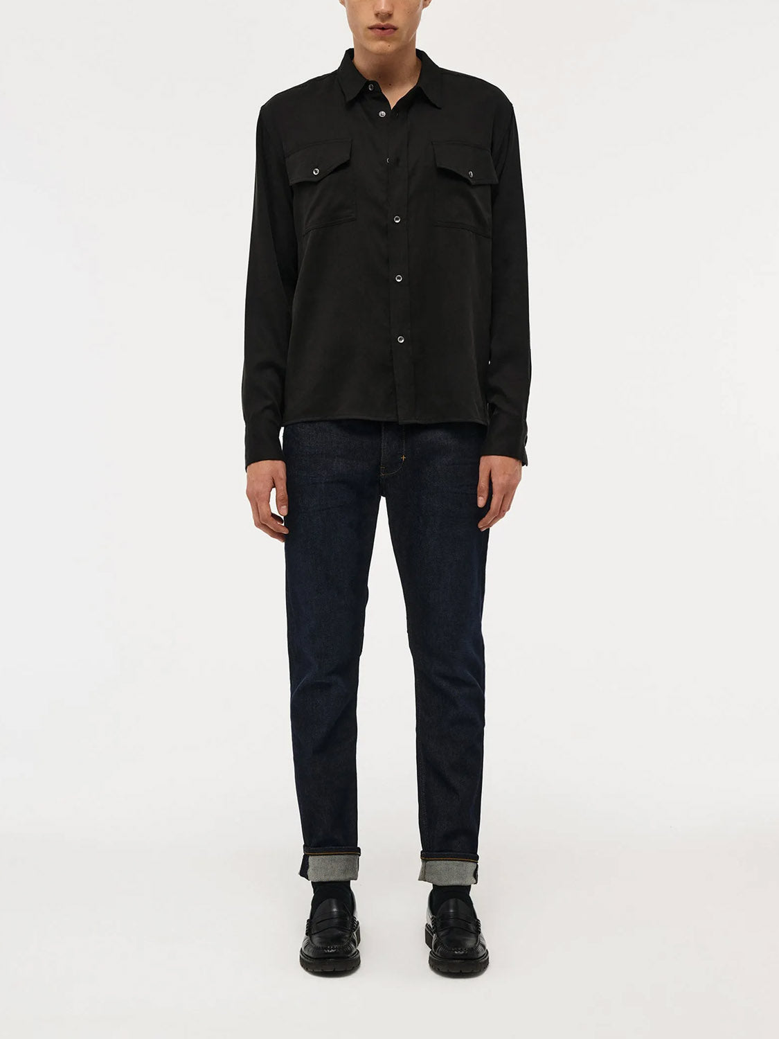 A person stands against a plain white background, wearing a black button-up shirt with chest pockets, Lou Slim - Typecast dark blue jeans by ID Menswear cuffed at the ankles, and black shoes. The person's head is partially out of frame.