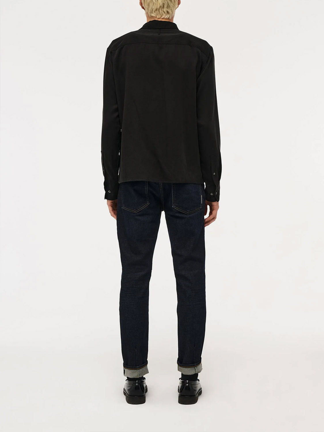 A person with short blond hair stands with their back to the camera, wearing a black long-sleeve shirt, ID Menswear Lou Slim - Typecast jeans with cuffs, and black shoes against a plain white background.