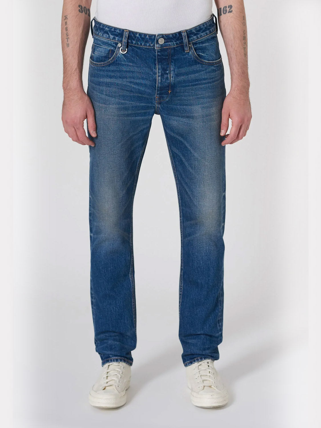 A person wearing Neuw Lou Slim Seventeen dark indigo straight-leg jeans, a white shirt, and white sneakers stands against a plain white background with relaxed arms, displaying tattoos on both forearms.