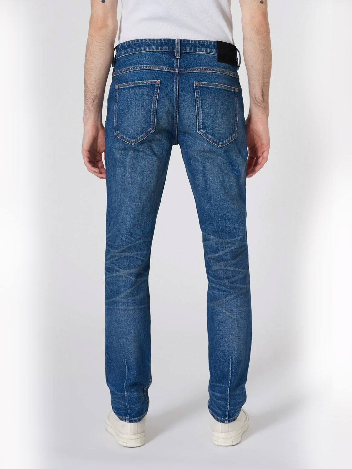 A person wearing Neuw’s Lou Slim Seventeen jeans in dark indigo, a white t-shirt, and white sneakers stands facing away from the camera. The slim fit jeans feature visible stitching and back pockets crafted from premium stretch denim.