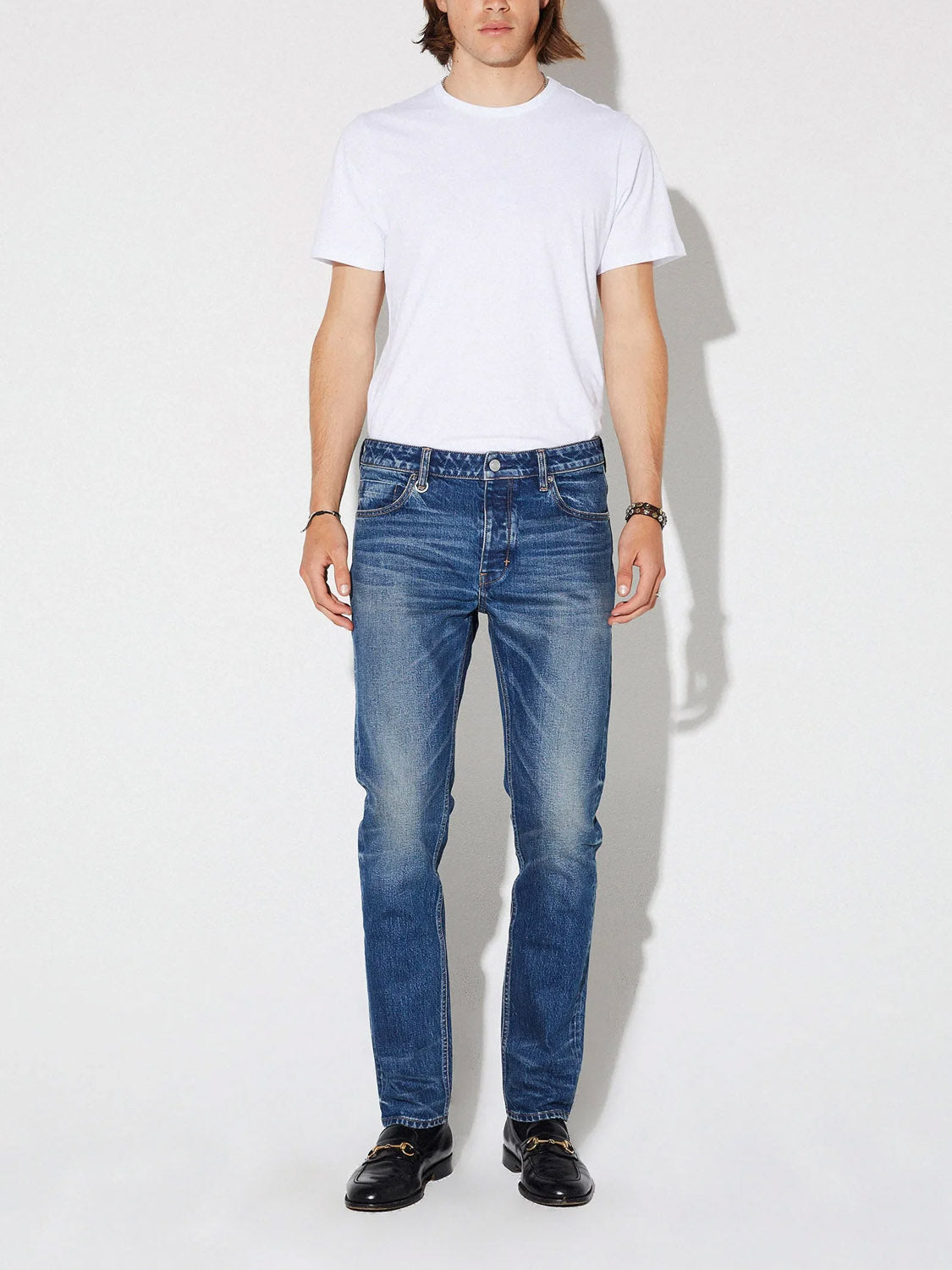 A person stands against a plain background, wearing a white short-sleeve t-shirt, Neuw Lou Slim Seventeen dark indigo jeans, black shoes, and a watch on their left wrist. Their face is partially out of frame.