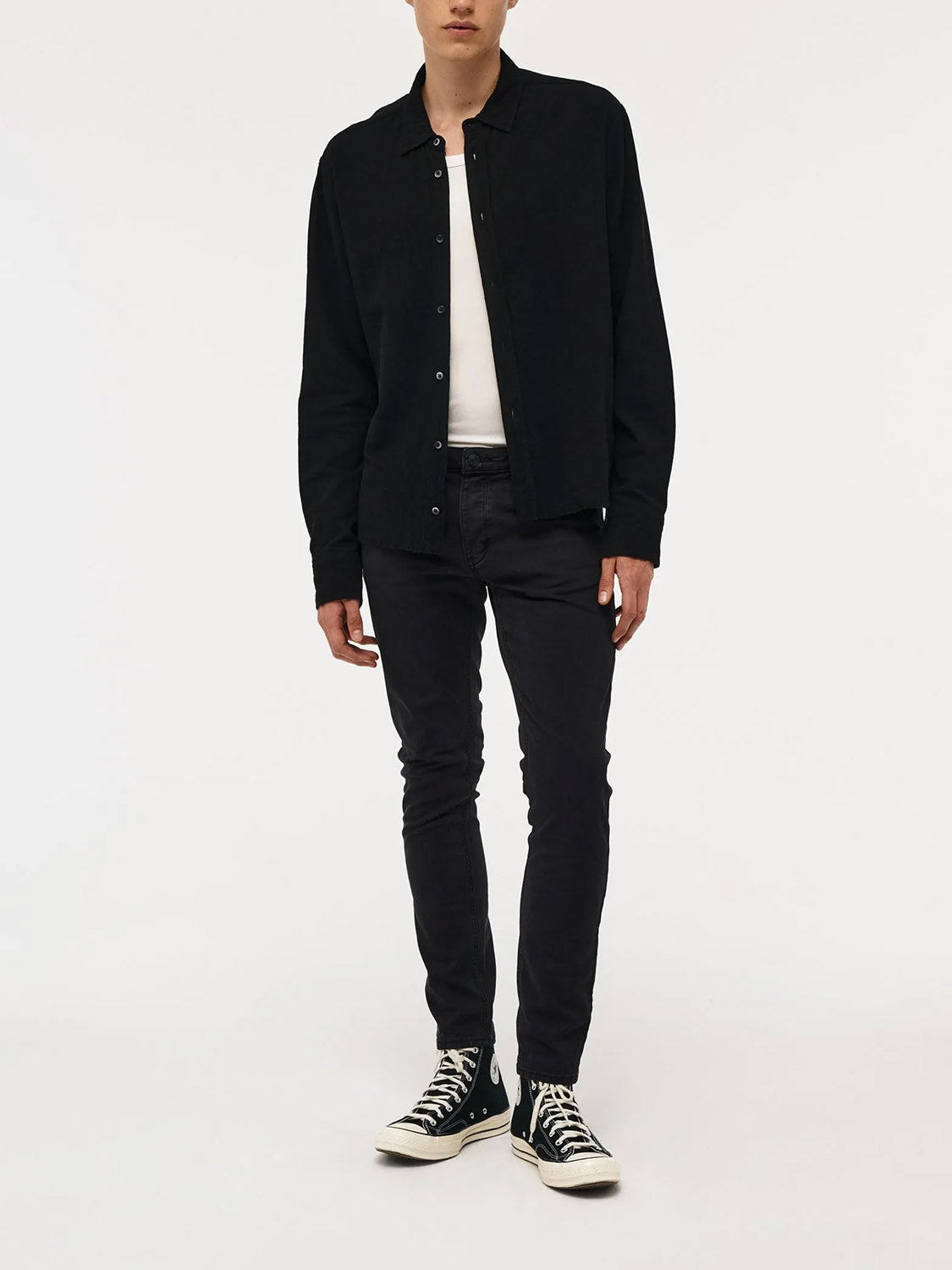 A person stands against a plain white background, wearing a black button-up shirt over a white tee, Neuw Lou Slim Twill Black jeans, and black high-top sneakers. Their hands are relaxed at their sides.