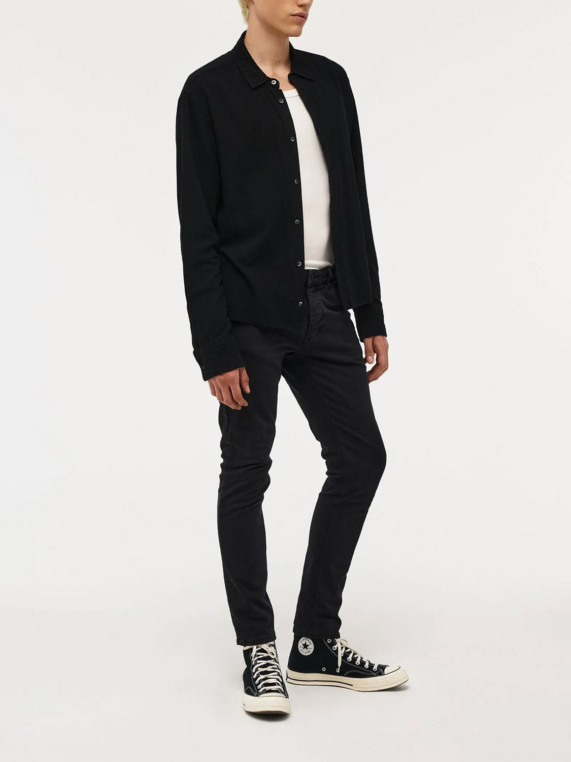 Against a white background, a person wears a black button-up shirt, white t-shirt, and Neuw Lou Slim Twill Black jeans. The look is finished with black high-top Converse sneakers. Their face is not visible.