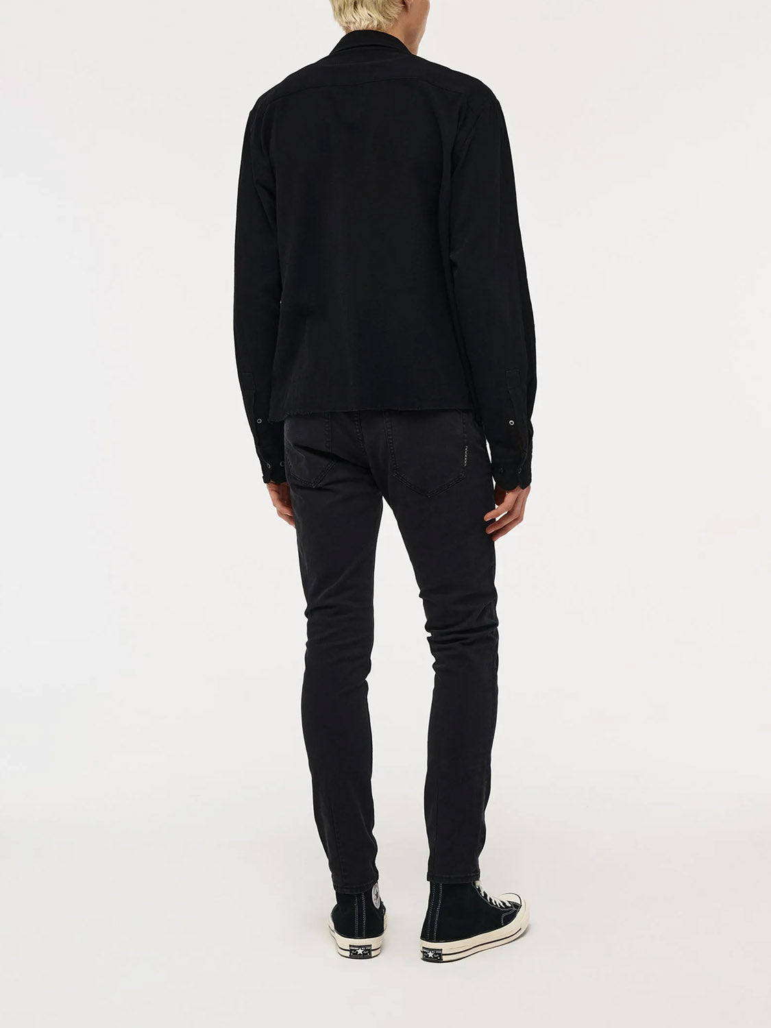 A person with short blond hair, dressed in a black jacket and Neuw Lou Slim Twill Black jeans, paired with black sneakers featuring white soles, stands facing away against a plain white background.