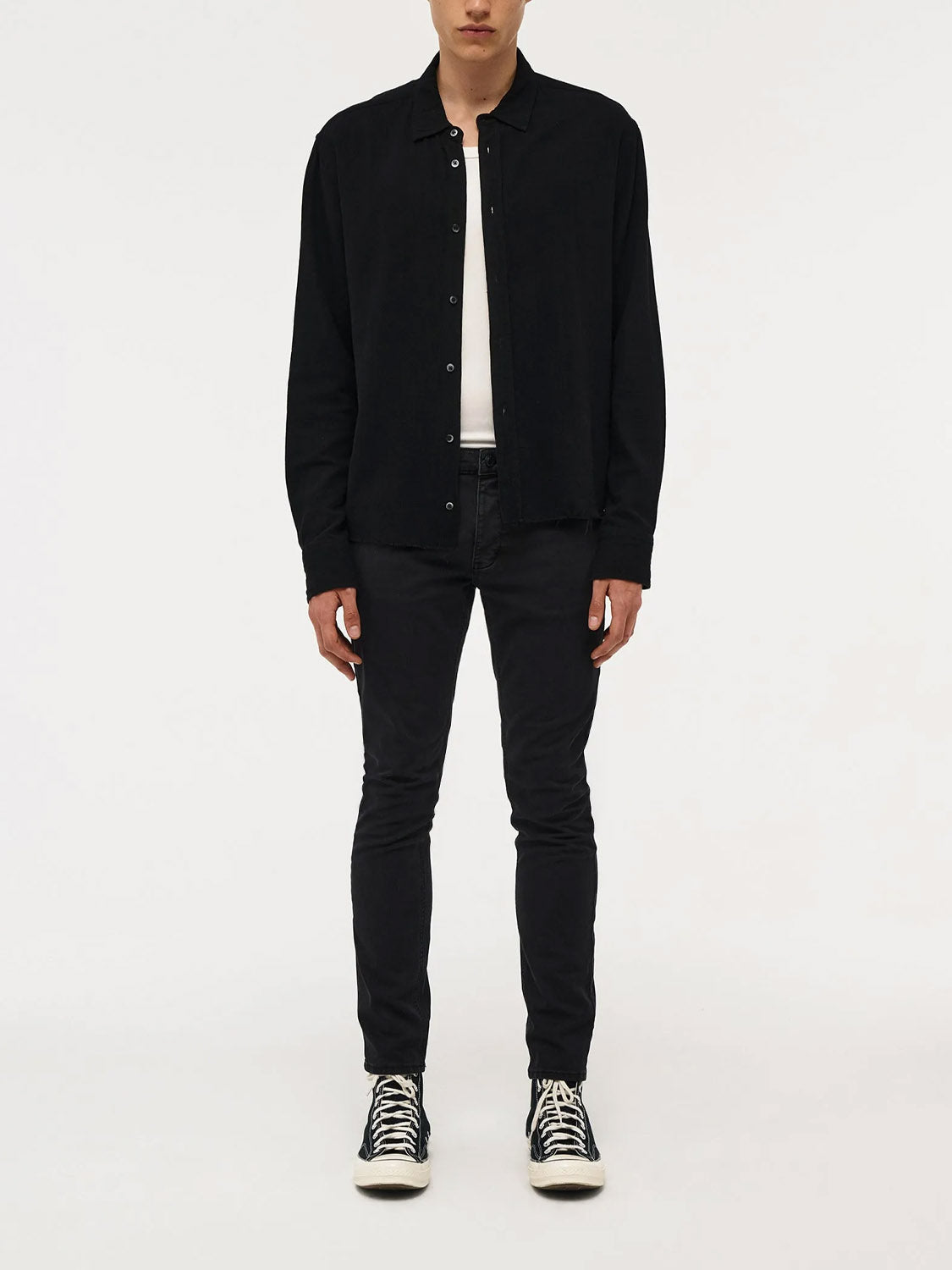 A person stands against a plain white background wearing a black button-up shirt, white t-shirt, Neuw Lou Slim Twill Black jeans, and black high-top sneakers with white laces and soles. Their hands are relaxed by their sides.