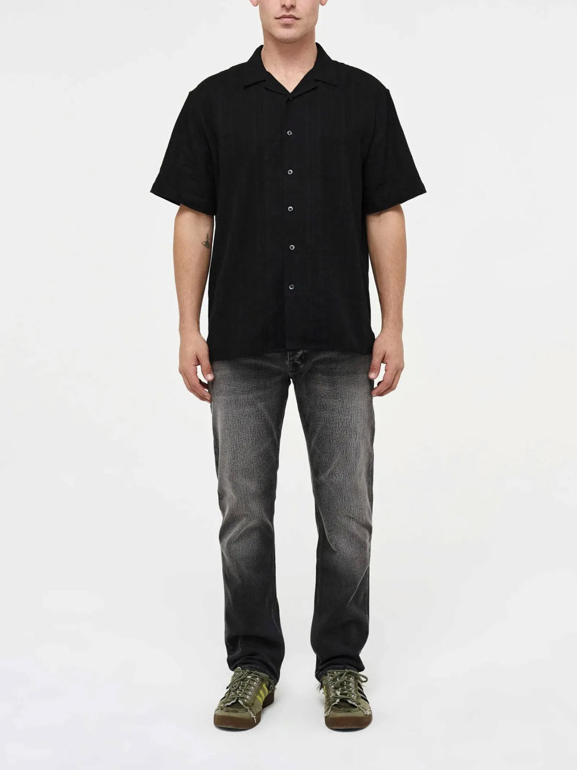 A man faces forward against a plain white background, wearing a black short-sleeve button-up shirt, Neuw Lou Straight Argent jeans in faded gray premium stretch denim, and green sneakers. His hands rest relaxed at his sides.