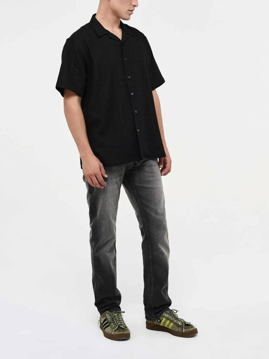 A person stands against a white background wearing a short-sleeved black button-up shirt, Neuw Lou Straight Argent premium stretch denim faded gray jeans, and green sneakers. Their face is turned slightly away from the camera.