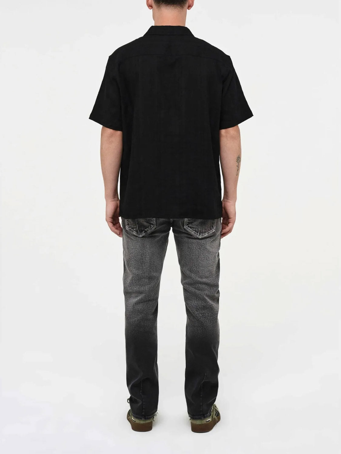 A person faces away from the camera, wearing a black short-sleeve shirt, Neuw Lou Straight Argent jeans in premium stretch denim, and brown shoes against a plain white background.