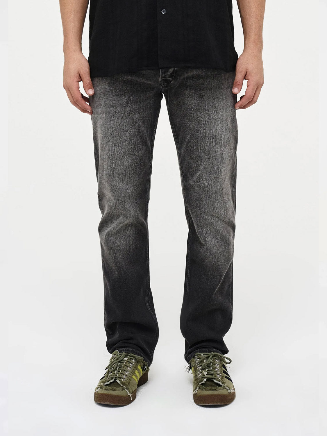A person wearing Neuw Lou Straight Argent faded black premium stretch denim jeans, a black shirt, and green sneakers stands facing forward against a plain white background. Photo is from the shoulders down.