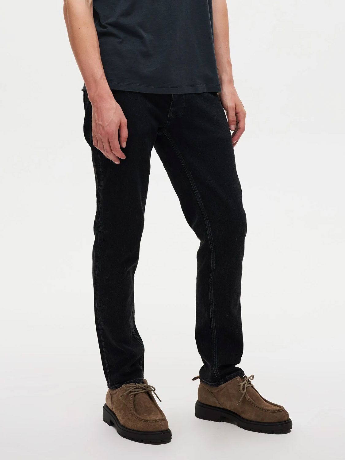 A person stands against a plain white background, shown from shoulders down, wearing a dark short-sleeve shirt and Neuw Lou Straight Dune jeans with brown suede lace-up shoes.