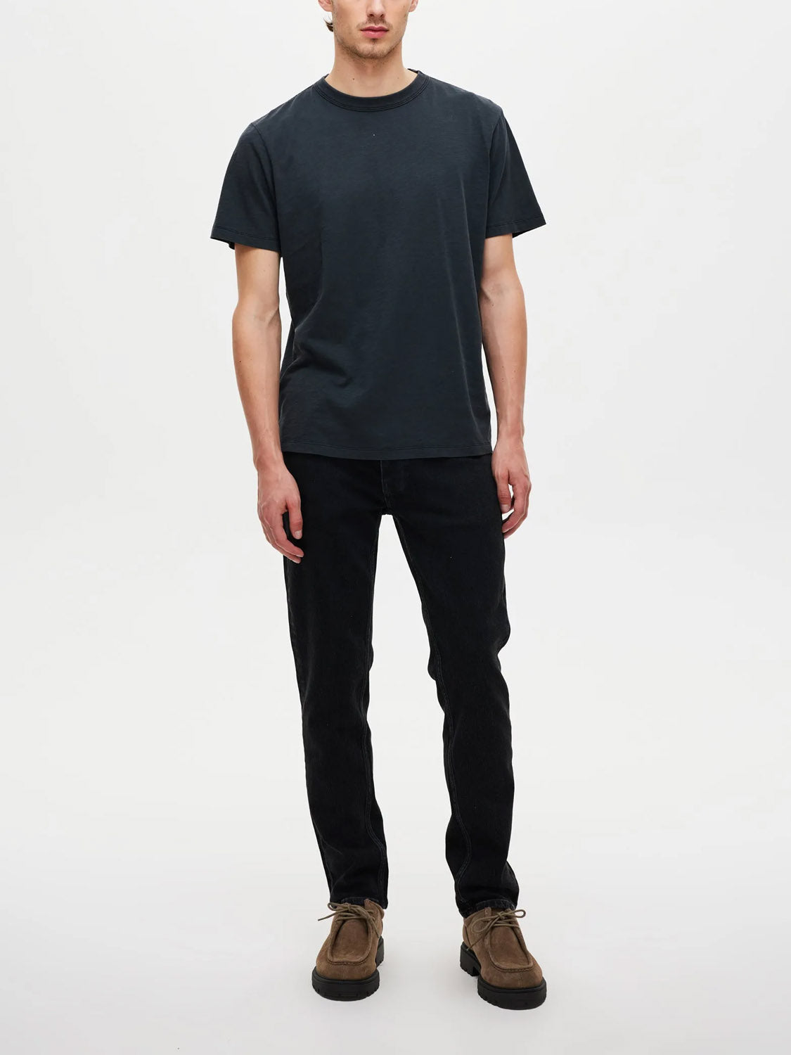 A person stands facing forward against a plain white background, wearing a plain black t-shirt, Neuw Lou Straight Dune jeans for a relaxed look, and brown shoes.