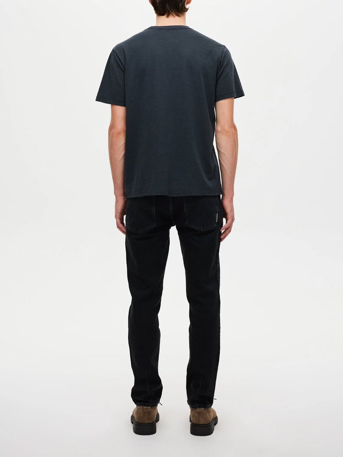 A person faces away from the camera, wearing a dark short-sleeve t-shirt, Neuw Lou Straight Dune jeans with a relaxed fit, and brown shoes, against a plain white background.