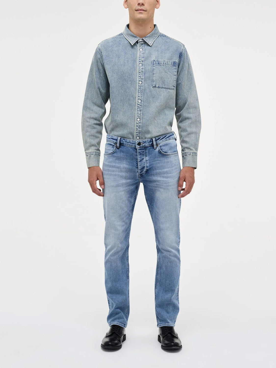 A man faces forward against a plain white background, wearing Neuw Lou Straight Fazer jeans with a light denim button-up shirt tucked in and black shoes.