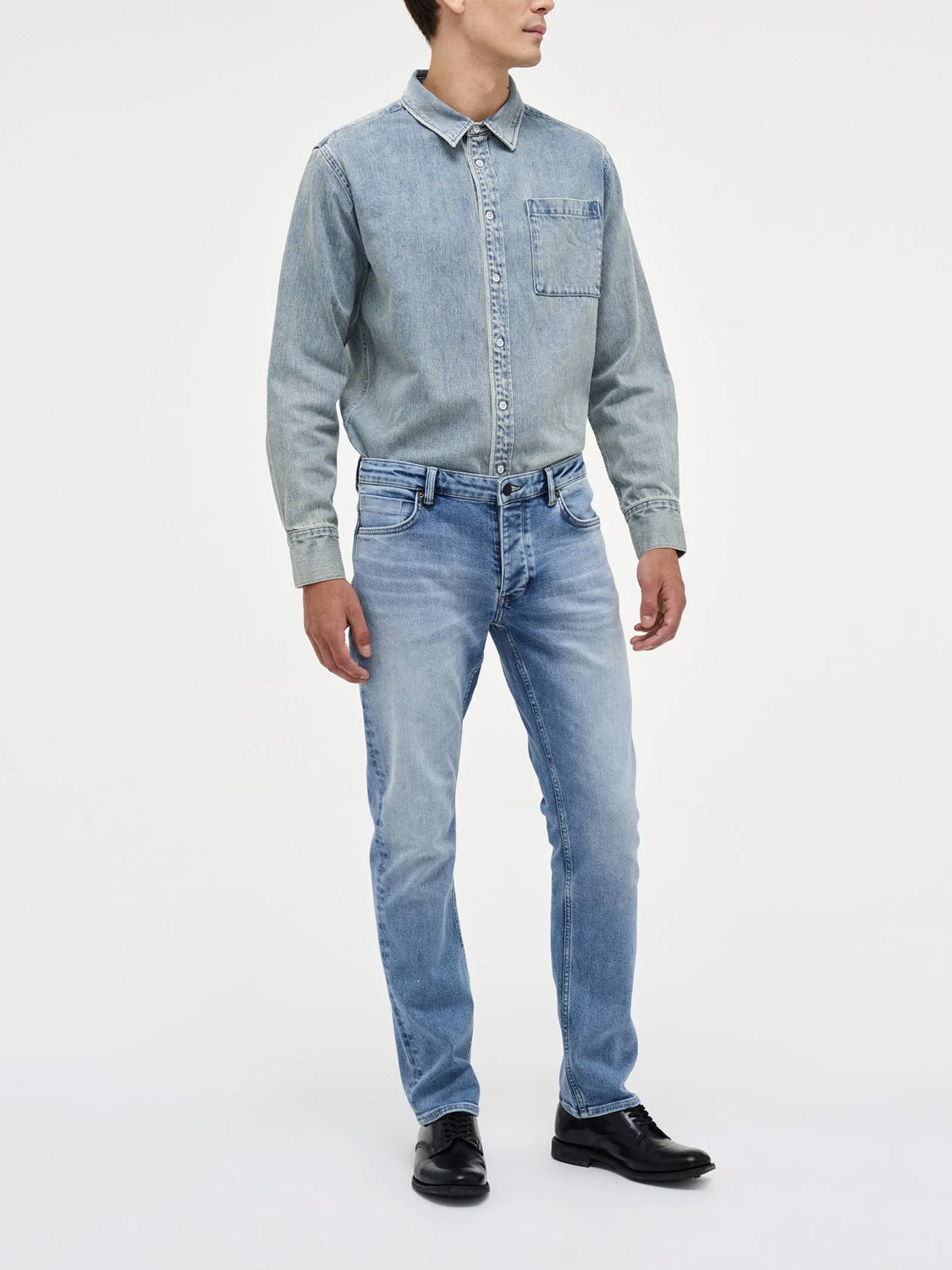 A person stands against a plain backdrop wearing Neuw’s Lou Straight Fazer light blue jeans, a relaxed denim shirt with a chest pocket buttoned up, and black shoes, with their head turned slightly to the side.