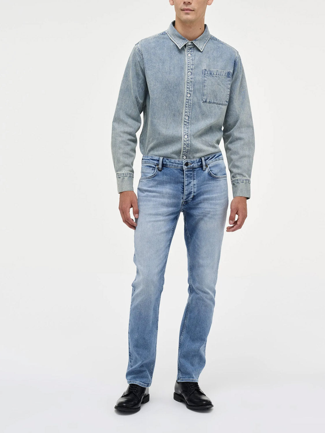 A person wears the Neuw Lou Straight Fazer jeans with a light blue denim shirt and black shoes, standing against a plain white background. The face is not visible.