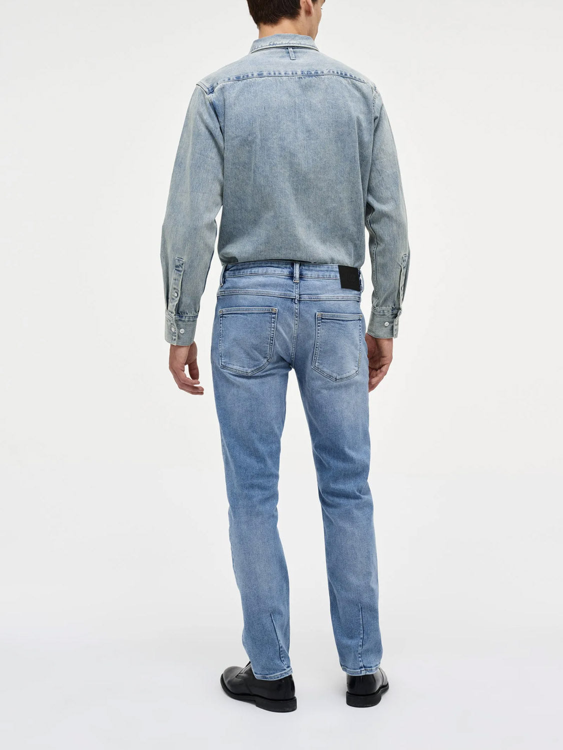 A man faces away from the camera, wearing Neuw Lou Straight Fazer jeans, a light blue denim shirt, and black shoes, presenting a relaxed silhouette against a plain white background.