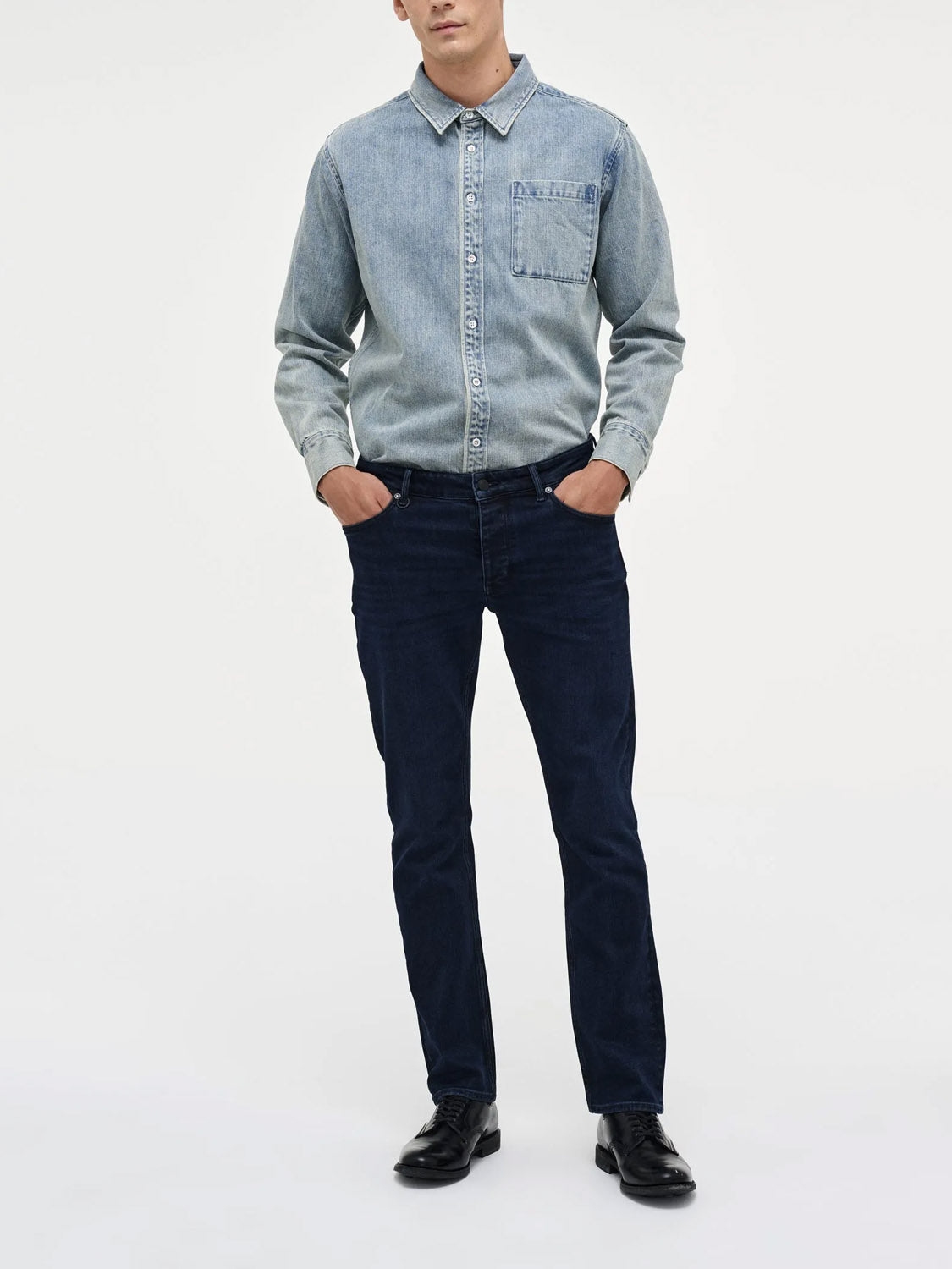 A person stands with hands in pockets, wearing Neuw’s Lou Straight Silent Water jeans and a light blue denim shirt with black shoes against a plain white background.