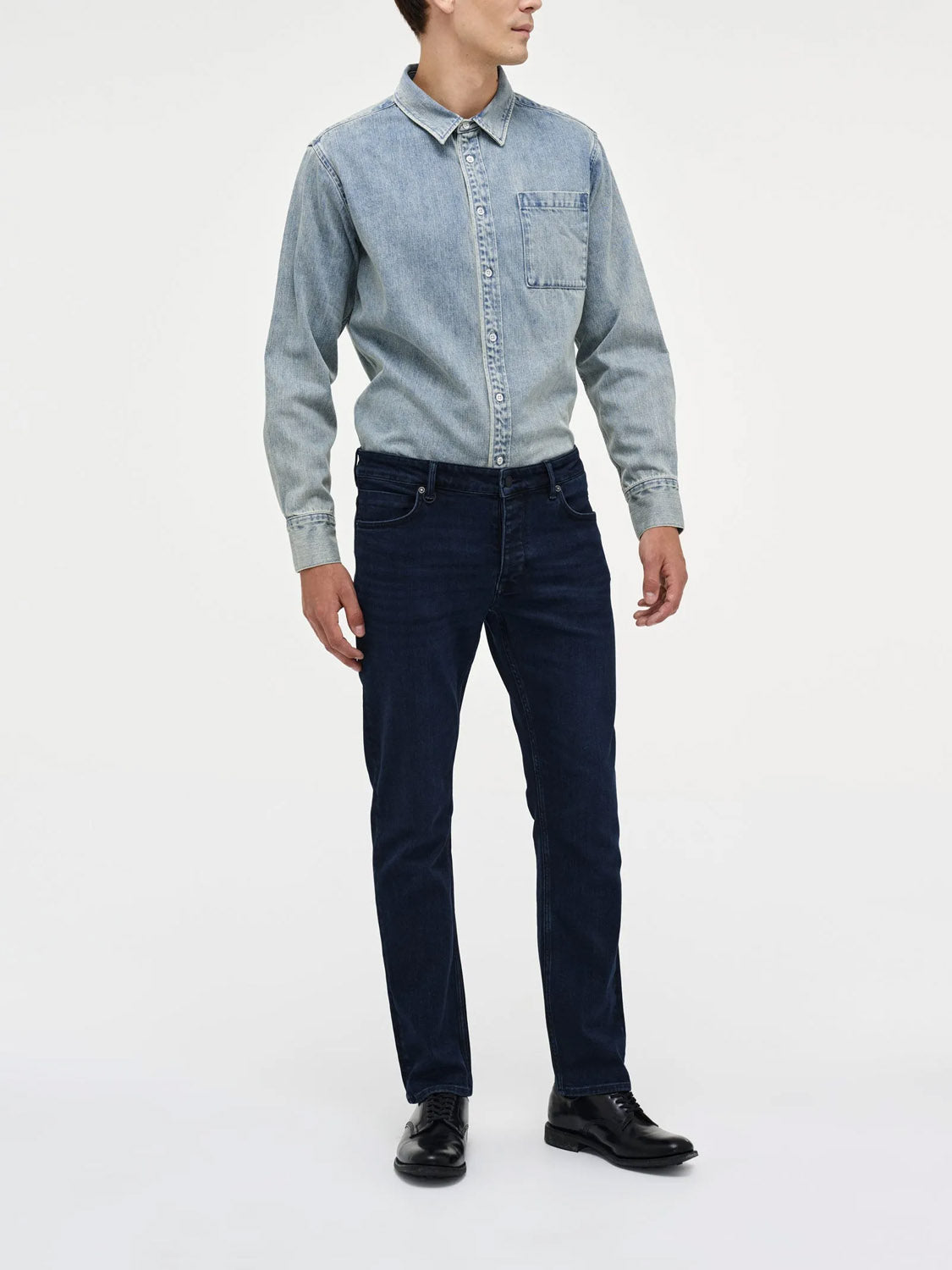 A person stands against a white background wearing Neuw Lou Straight Silent Water indigo denim jeans, a light blue denim button-up shirt, and black dress shoes.