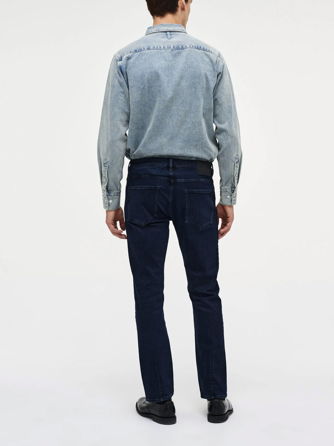 A man stands with his back to the camera, wearing Neuw's Lou Straight Silent Water indigo denim jeans and a light blue shirt, set against a plain light background.