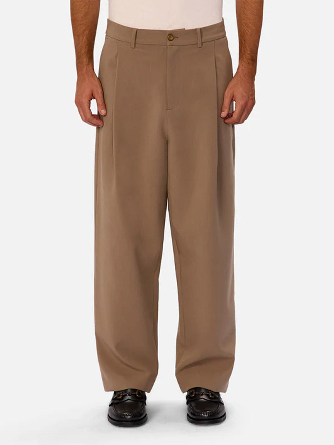 A person wearing Industrie’s Lusso Loose Fit Pant in tan, paired with a tucked-in white shirt and black dress shoes, stands against a plain light background. Only the lower torso and legs are visible.
