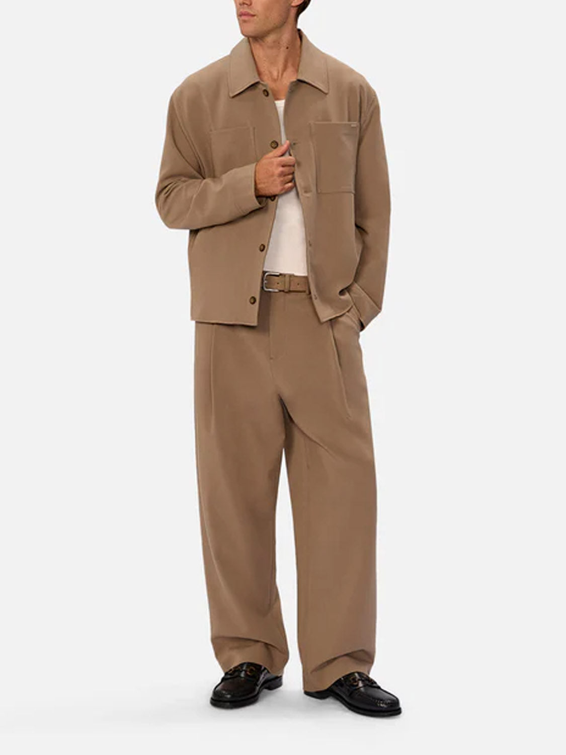 A man stands against a light background, one hand in his pocket and the other adjusting his jacket, dressed in Industrie’s Lusso Loose Fit Pant and a white shirt under a tan suit.