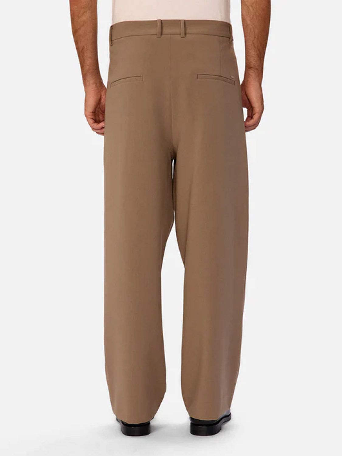 A person wearing Industrie Lusso Loose Fit Pant in brown stretch fabric, seen from the back with hands by their sides. Featuring two back welt pockets, the pants are styled with black shoes and a tucked-in shirt.