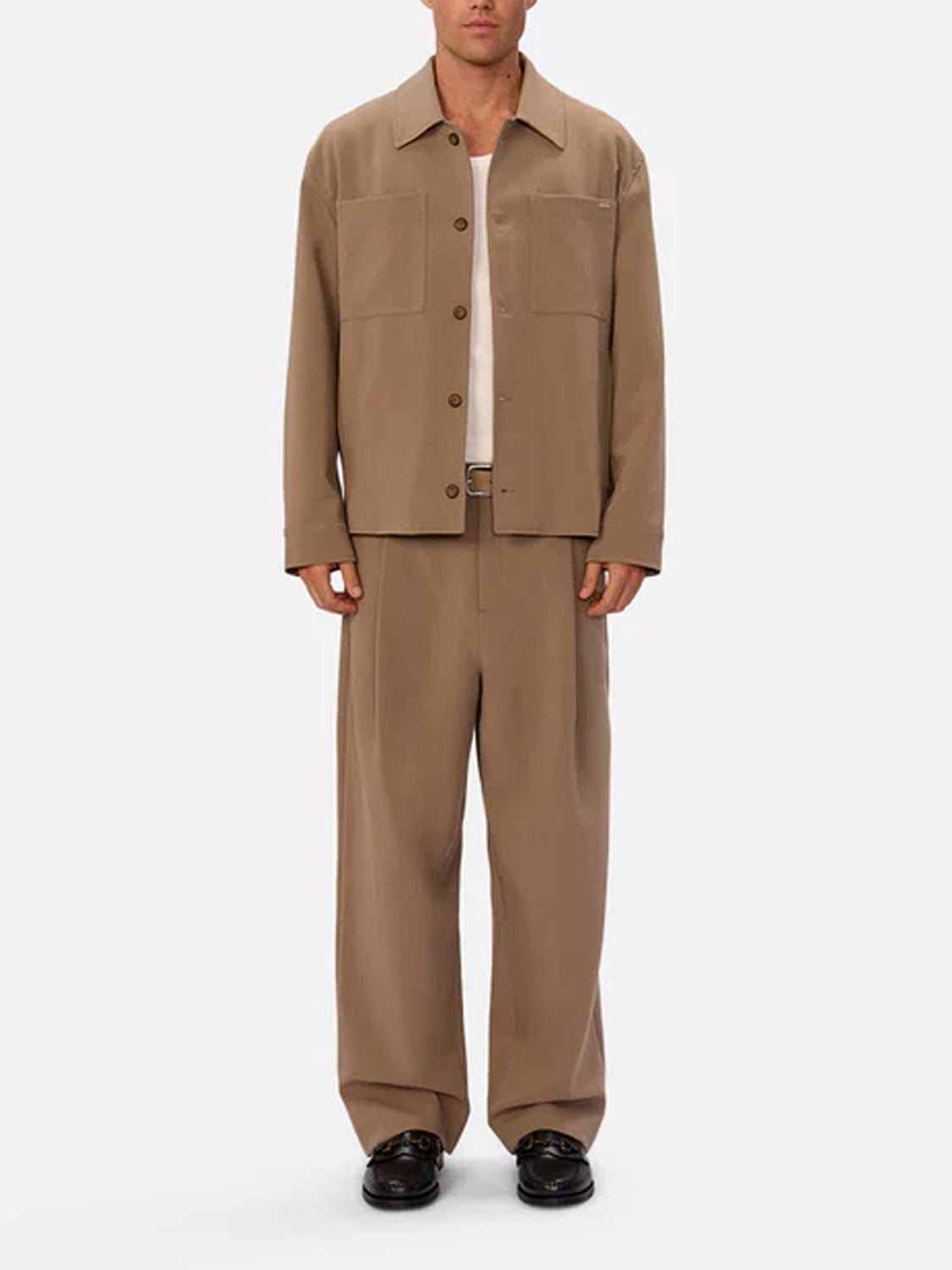 A man faces forward against a plain light background, wearing Industrie’s Lusso Loose Fit Pant and a loose tan jacket with large front pockets, paired with a white undershirt and black shoes.