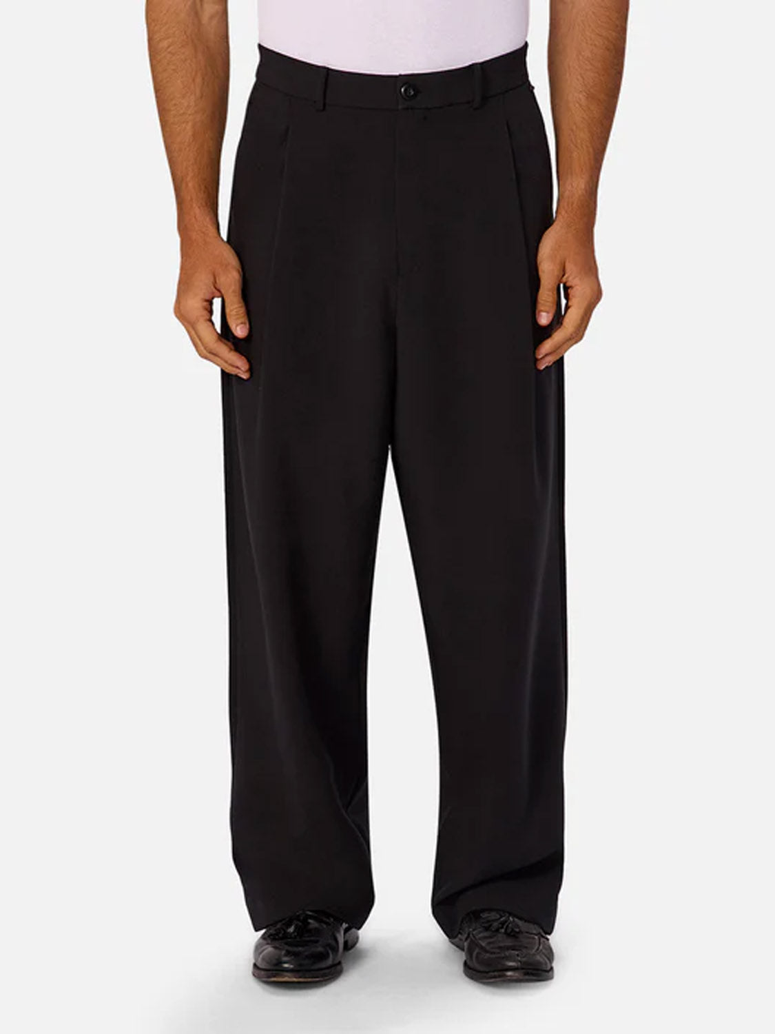 A person wearing Industrie’s Lusso Loose Fit Pant in black stretch fabric, paired with a tucked-in white shirt and black dress shoes, stands against a plain light gray background.