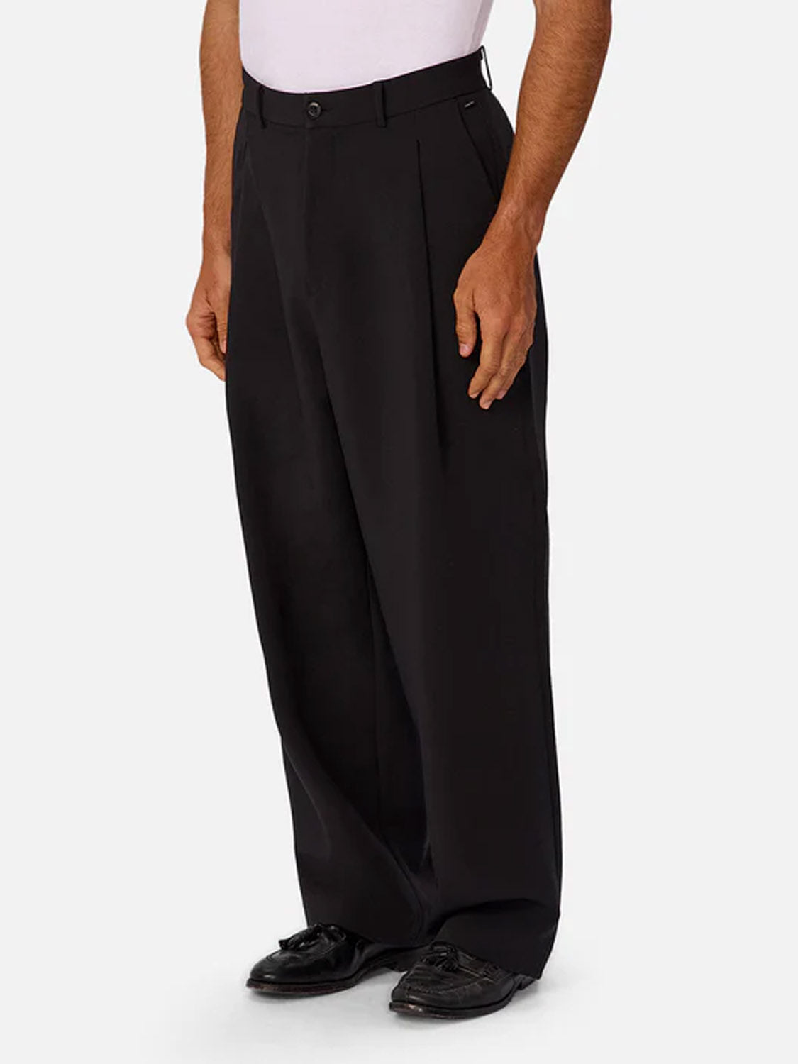 A person wears Industrie’s Lusso Loose Fit Pant in black with a tucked white shirt and black dress shoes, standing against a plain light gray background, shown from shoulders down.