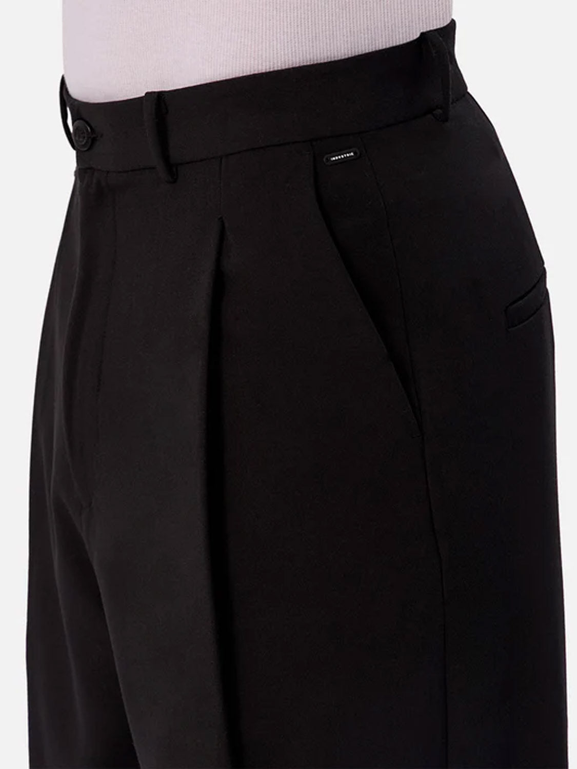 Close-up of a person wearing the Lusso Loose Fit Pant by Industrie, featuring a high waist, black relaxed fit, pleats, belt loops, button closure, and side pocket. The upper part of a light-colored top is partially visible.