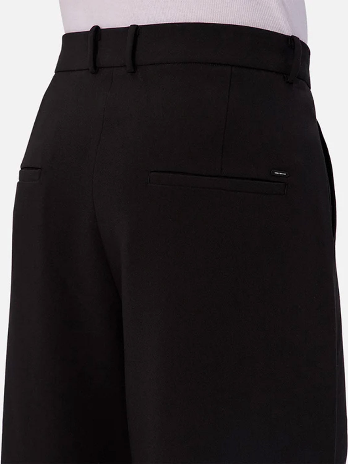 Back view of a person wearing Industrie Lusso Loose Fit Pant in black stretch fabric with belt loops and two back welt pockets, one accented with a metallic brand tag. The upper part of a tucked-in white shirt is visible.