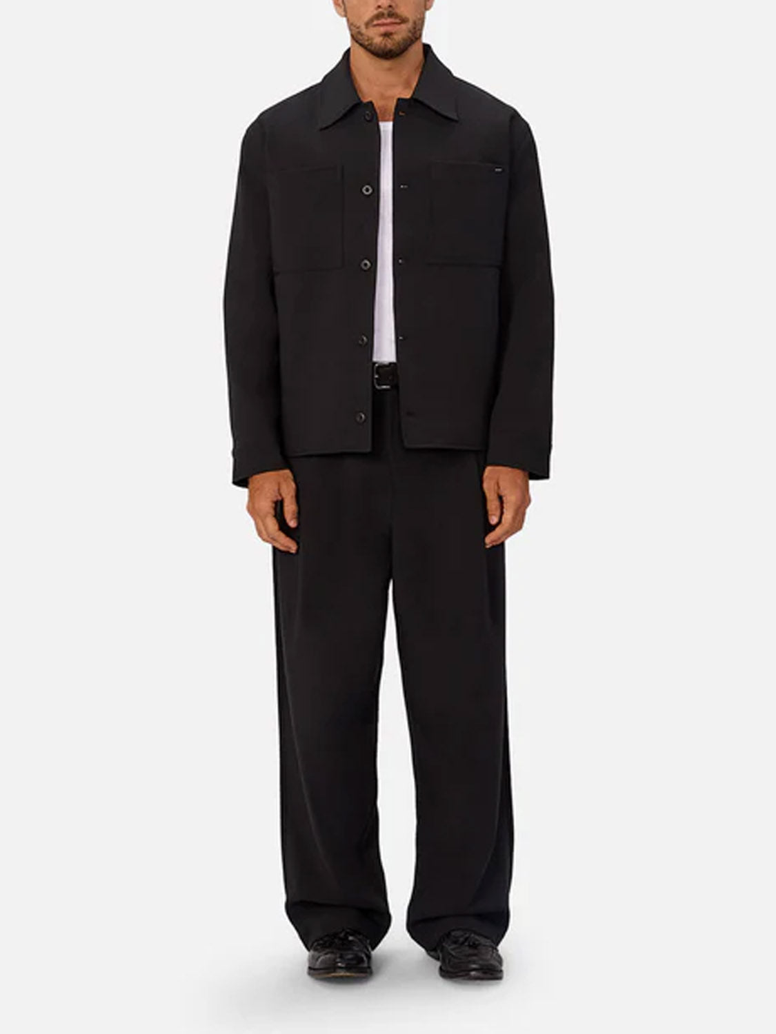 A man stands facing forward, wearing a black button-up jacket, white t-shirt, Industrie Lusso Loose Fit Pant, and black shoes against a plain light gray background.