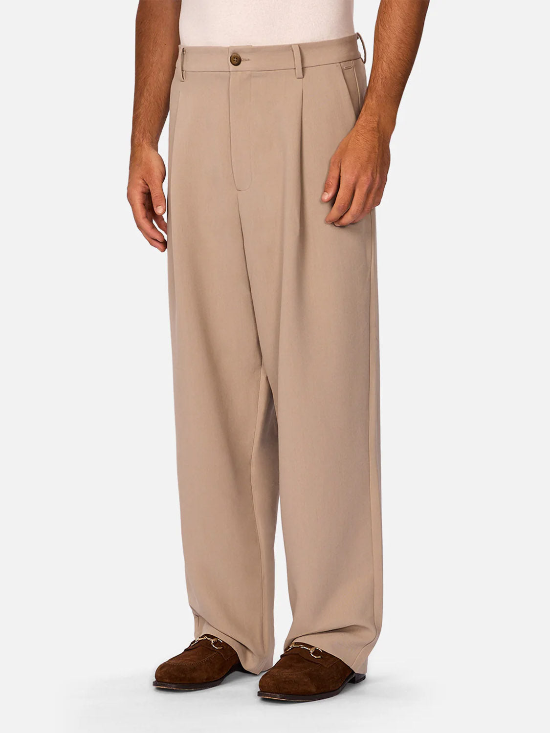 A person wears Industrie’s Lightweight Classic Lusso Pant - Stone, featuring pleats and belt loops, paired with brown loafers, standing against a plain light background. Only the lower half of the body is visible.